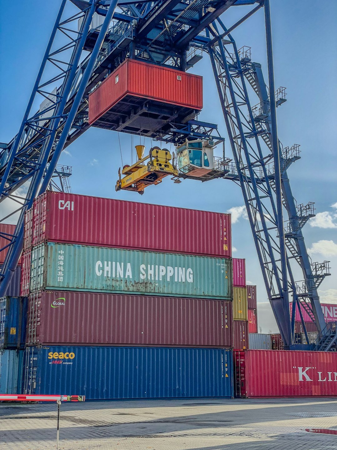A gantry crane lifting a red shipping container above a stack of various containers with CHINA SHIPPING visibly marked, under a blue sky with clouds.